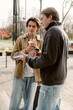 © m-art - Two young men stand outside, holding drinks and engaged in conversation. The sun shines brightly on a cheerful day, and trees surround them, creating a relaxed atmosphere.