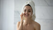 © fizkes - Headshot portrait cheerful young woman look at mirror after morning water procedures with hair wrapped in towel take care of face skin apply cosmetic cream mask serum lotion for hydration rejuvenation