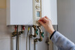 © Alina Mosinyan - Hand turning the control knob on a white wall-mounted gas boiler, with pipes visible below, representing residential heating systems and everyday energy management at home.