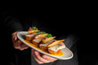 © pavel siamionov - waiter holding a grey oval plate with several pieces of elegant fish canapés or appetizers garnished with pear slices, red onion, and parsley on a yellow sauce, against a black background