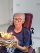 © Creativa Images - Smiling Elderly Man in Wheelchair Holding a Birthday Cake with Lit Candles