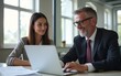 © David - 2 smiling executives using laptop discussing work. Busy colleagues talking on project strategy at meeting table. Two professional middle aged business man and woman managers in office. Authentic photo