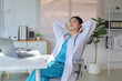 © amnaj - Asian female doctor in scrubs and lab coat, leaning back in an office chair, eyes closed, enjoying a relaxing moment during a break
