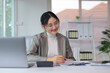 © amnaj - Asian businesswoman analyzing data and writing notes on documents at her desk in a modern office, smiling
