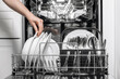 © wedmoments.stock - Hand placing clean white plates into a modern dishwasher with stainless steel utensils, showcasing efficient kitchen appliance use and organized dishware storage