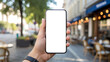 © Art_spiral - First person point of view of a hand holding a smartphone with a blank white mockup screen on a sunny city street with a blurred outdoor cafe and sidewalk terrace in the background
