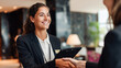 © Andrey - Smiling female hotel receptionist in black blazer shaking hands with a guest, holding a digital tablet, professional hospitality service in a luxury lobby, warm indoor lighting.