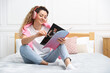 © New Africa - Happy woman in headphones with magazine on bed near white wall at home