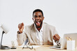 © SHOTPRIME STUDIO - Happy young African American man in beige blazer celebrates success at office desk, smiling and raising fist, surrounded by office supplies, cheerful mood, professional attire, upbeat atmosphere