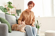 © Pixel-Shot - Mature redhead woman with cute Maine Coon cat sitting on sofa at home