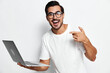 © SHOTPRIME STUDIO - Smiling man wearing white t-shirt holding laptop while pointing finger at camera with cheerful expression, isolated on plain solid background. Tech lifestyle concept.