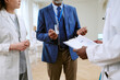 © AnnaStills - Middle aged Black man in suit discussing medical report with young adult Caucasian woman and young adult Black man in lab coats, standing together in hospital setting