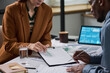 © pressmaster - Caucasian young adult woman and Black young adult man reviewing financial documents together at desk, both focusing on paperwork with digital tablet displaying charts in background
