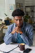 © pressmaster - Young adult Black man sitting at desk contemplating, paperwork and coffee mug on table, smartphone nearby, office shelves and window in background