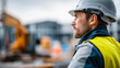 © Denis Yevtekhov - Faceless civil engineer in reflective jacket with safety helmet looks into distance at construction site with excavator in background, site supervision, project oversight, defocused person, with