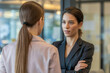 © Igor - Two professional business women having a serious conversation in a modern corporate office hallway with one woman wearing a dark blazer and crossing her arms during a meeting.