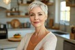 © Tatyana - Confident middle-aged woman with short gray hair standing in a sunlit modern kitchen with arms crossed, calm and content in a cozy home interior