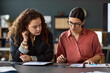 © DragonImages - Caucasian young adult woman and Hispanic young adult woman collaborating at desk, reviewing documents and using digital tablet, both focused on paperwork during business meeting