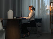© kinomaster - A professional woman wearing headphones, working on a laptop at a table near a window, illuminated by warm light.