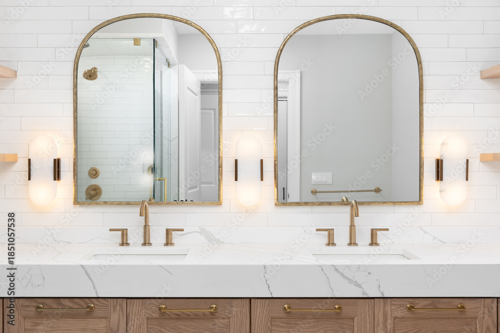 A bathroom detail with gold faucets, light fixtures, and mirrors, a wood cabinet, large marble slab countertop, and a subway tile wall.