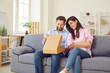 © Studio Romantic - Young happy couple opening cardboard box sitting on sofa in living room at home. Man and woman buyers unpacking parcel receiving online store order. Delivery and shopping online concept.