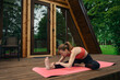 © boomeart - Woman practices yoga on a pink mat outside a wooden cabin in the morning