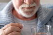 © luciano - An elderly man with flu or COVID symptoms takes a pill with a glass of water. A stressed, bearded older man takes antidepressants or sedatives. Medications at work.