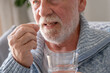 © luciano - Senior man takes pill with glass of water in hand. Stressed elderly bearded man drinking sedated antidepressant meds. Man feels depressed, taking drugs. Medicines at work
