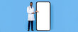 © Prostock-studio - A healthcare professional in a white coat stands next to a large blank phone screen. He is smiling and gestures with his hand, suggesting an innovative approach to health technology.