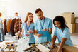 © Prostock-studio - Young arab male activist checks a donation list while other volunteers pack boxes with food and supplies. The scene shows teamwork and community support in a charitable organization setting.