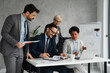 © Novak - Four diverse business professionals working together at a large desk in a bright, modern office, collaborating on a digital project.