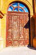 © Leilani - Weathered red wooden door with peeling paint stands within yellow stone archway, topped by segmented transom window. Aged texture and rustic charm evoke history, exposure and architectural character