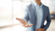 © sa-photo - Businessman in blue suit extending hand forward with blurred background, offering help or handshake in bright office environment