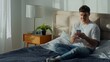 © Yuliia - Happy Caucasian man male sitting on bed at home smiling using smartphone young guy holding mobile phone texting online chat browsing internet sending message relax gen z leisure weekend comfort indoor