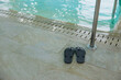© True Pixel Art - A pair of dark flip flops rests on wet stone tiles by a stainless handrail and slot drains, rippling turquoise water reflects soft indoor light, low angle, shallow depth.