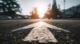 Close-up asphalt road, leading arrow with bright sun, trees and blurred buildings