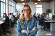 © Aleksei Solovev - A smiling woman with glasses and a denim jacket stands confidently in a busy, modern office space.