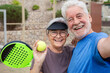 © Daniel - Old senior couple outdoors in paddle tennis court smiling and looking at the camera taking a selfie picture together playing padel. Active mature people concept