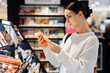 © wedmoments.stock - Young woman examining colorful cosmetics in a beauty store, surrounded by various makeup products on display, showcasing her interest in beauty and self-care