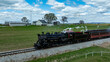 © Greg Kelton - A steam train moves on rails through an open field with a farmhouse in the background. Smoke rises from the engine as the train progresses under a blue sky filled with clouds.