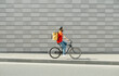 © Prostock-studio - Deliveryman on bicycle. Young guy with beard and big backpack rides along path in city on gray brick wall background, side view, free space