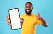 © Prostock-studio - New cool mobile app. Funky African American guy demonstrating cellphone with empty screen, showing thumb up gesture, recommending website, offering space for advertisement on blue background, mockup