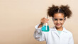 © Mariya Sorvacheva - Young girl in a white lab coat and safety goggles holds a flask with blue liquid, showcasing her enthusiasm for science in a bright classroom setting