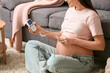 © Pixel-Shot - Young pregnant woman with fetal doppler listening to baby's heartbeat near sofa at home, closeup