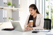 © Fahng - Young business woman  holding cup drinking coffee working on laptop computer sit alone at table in office indoors .