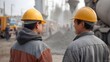 © Thares2020 - Two construction workers in yellow hard hats converse at a dusty industrial site with hinery in the background