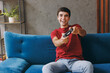 © ViDi Studio - Young excited man wear red t-shirt hold play pc game with joystick console sit on blue sofa couch stay at home hotel flat rest relax spend free spare time in living room indoors People lounge concept.