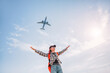 © EdNurg - Young woman with backpack cheerfully raising arms while a passenger jet flies overhead, symbolizing travel, freedom, and vacation