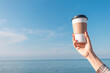 © EdNurg - Hand holding a takeaway coffee cup against a clear blue sky and calm ocean, symbolizing relaxation and vacation