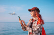© EdNurg - Young woman traveler using smartphone on beach, smiling while a passenger airplane approaches for landing in background